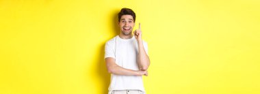 Çekici bir adamın fikri olduğunu hayal etmek, parmağını kaldırıp plan önermek, heyecanlı gülümsemek, sarı arka planda dikilmek..