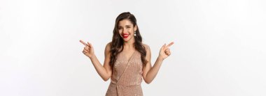 Parti zamanı. Zarif elbiseli güzel bir kadın, kırmızı dudaklı, parmaklarını yana doğru gösteren, sol ve sağ promosyon tekliflerini gösteren, gülümseyen ve kameraya bakan, beyaz arka plan.