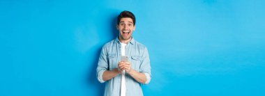 Şaşırmış ve mutlu bir adam internette bir şeyler kazanıyor, elinde akıllı telefon ve sevinçle, mavi arka planda duruyor..