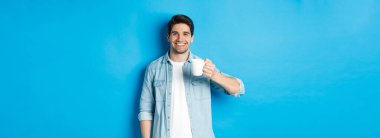 Elinde kupa tutan ve kahve içen, mavi arka planda duran gülümseyen sakallı adam..