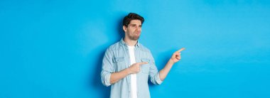 Şüpheli yetişkin bir adam terfiyi sol eliyle işaret ediyor ve kararsız görünüyor, hayal kırıklığına uğruyor, mavi arka planda duruyor..
