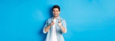 Küstah yakışıklı adam seni işaret ediyor, flört ediyor, mavi arka planda sıradan kıyafetler içinde dikiliyor..