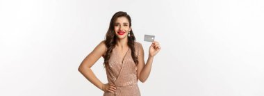 Tatillerde. Noel partisi ve alışveriş konsepti. Kırmızı dudaklı şık bir kadın, gösterişli bir elbise, kredi kartı gösteriyor ve gülümsüyor, beyaz arka planda duruyor..