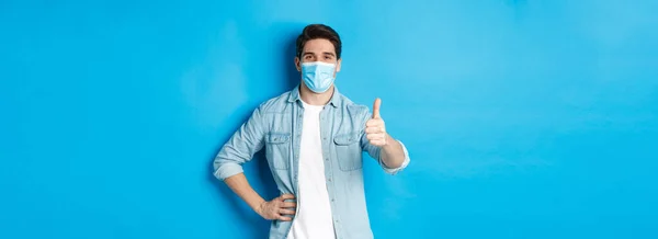 Concept of covid-19, pandemic and social distancing. Satisfied guy in medical mask showing thumb up in approval, standing against blue background.