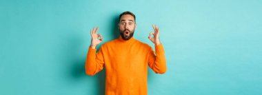 Etkileyici bir adam süper havalı bir şey övüyor, tamam işareti gösteriyor ve kameraya bakıyor, açık mavi arka plan.