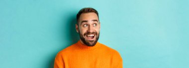 Yakışıklı, beyaz bir adam gülümsüyor, şaşırmış bir şekilde sola bakıyor, logoya bakıyor, turuncu süveter giyiyor, turkuaz arka planda duruyor..