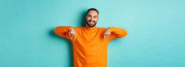 Mutlu, sakallı, parmaklarını aşağı gösteren, neşeli gülümseyen, reklam veren, turkuaz arka planda duran yetişkin bir adam..