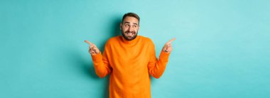 Kafası karışmış yakışıklı adam kararsız, parmaklarını yana doğru uzatıyor, iki değişken arasında seçim yapamıyor, açık mavi arkaplana karşı duruyor..