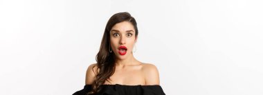 Moda ve güzellik konsepti. Siyah elbiseli çekici esmer kadının yakın çekimi ağzı açık, kameraya hayranlıkla bakıyor, beyaz arka plan.