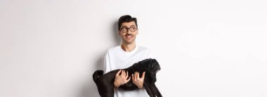 Köpeğiyle logoya bakan heyecanlı hippi adam. Yakışıklı evcil hayvan sahibi siyah pug taşıyor ve beyaz arka planda duran reklam panosuna bakıyor..