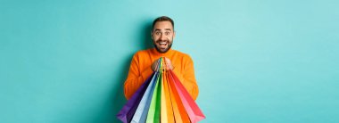 Alışveriş torbaları tutan, gülümseyen, alışveriş merkezine giden, turkuaz arka planda duran heyecanlı yetişkin bir adam..
