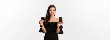 İnternetten alışveriş konsepti. Mobil ekran ve kredi kartı gösteren siyah elbiseli şık esmer kadın, gülümsüyor, beyaz arka planda duruyor..