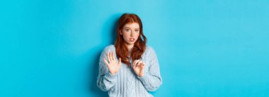 Endişeli kızıl kız teklifi reddediyor ya da reddediyor, el sıkışıyor ve kameraya endişeyle bakıyor, tatsız bir şeyi reddediyor, mavi arka planda duruyor..