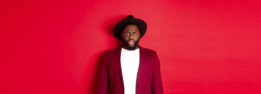Moda ve parti konsepti. Şık giyinmiş kafası karışmış siyah bir adam, kaşlarını çatmış, kameraya bakıyor, hoşnutsuz, anlayamıyorum, kırmızı arka planda dikiliyor..