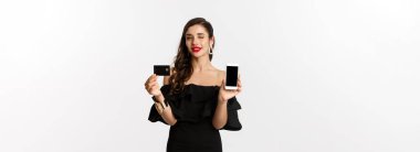 Moda ve alışveriş konsepti. Kırmızı dudaklı güzel bir kadın, kameraya göz kırpıyor, akıllı telefon ekranı ve kredi kartı gösteriyor, internetten alıyor, beyaz arka plan.