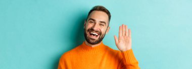 Dost canlısı genç adamın yakın çekimi gülümseyen, merhaba demek için el sallayan, seni selamlayan, açık mavi arka planda duran.