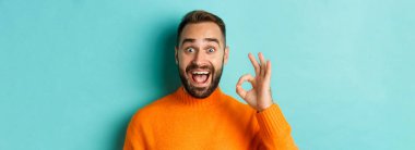Etkilenmiş yakışıklı bir adam iyi bir işaret veriyor, iyi bir şey öneriyor, iyi bir jestle övüyor, açık mavi arka planda duruyor..
