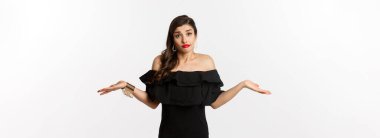 Moda ve güzellik. Siyah elbiseli cahil ve çekici bir kadın. Bilmiyor, omuz silkiyor ve sırıtıyor. Beyaz arka planda kararsız duruyor..