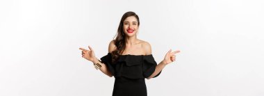 Moda ve güzellik. Mücevher, makyaj ve siyah elbise giymiş çekici bir kadın, gülümseyen ve parmaklarını yana doğru gösteren fotokopi alanı teklifi, beyaz arka plan.