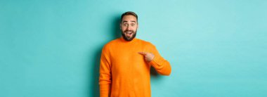 Heyecanlı adam kendini işaret ediyor, şaşkın ve mutlu görünüyor, seçiliyor, açık mavi arka planda duruyor..
