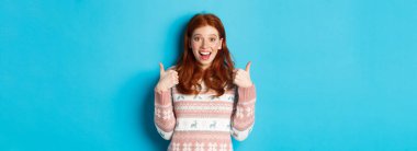 Kız harika bir şeyi onaylıyor, baş parmağını kaldırıyor, aferin sana ve gülümsüyor mavi arka planda duruyor..