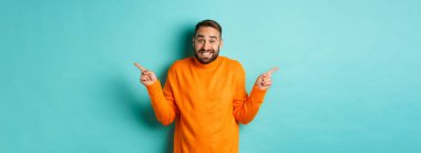 Kafası karışmış yakışıklı adam kararsız, parmaklarını yana doğru uzatıyor, iki değişken arasında seçim yapamıyor, açık mavi arkaplana karşı duruyor..