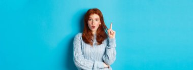 Düşünceli kızıl saçlı kızın bir fikri var, parmağını kaldırıyor ve önerilerde bulunuyor, mavi arka planda duruyor..