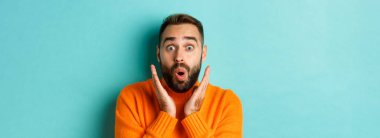 Kameraya bakan, turuncu süveter giyen, turkuaz arka planda duran yakışıklı beyaz adamın yakın çekimi..