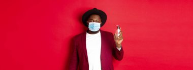 Covid-19, karantina ve tatil konsepti. Tıbbi maske takan ve el dezenfektanı gösteren şık giyimli genç Afro-Amerikan adam antiseptik kullanmayı öneriyor..