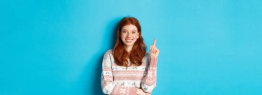 Heyecanlı genç kızın bir fikri var, parmağını kaldırıyor ve gülümsüyor, bir şeyler ima ediyor, mavi arka planda süveter giyiyor..