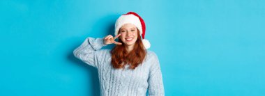 Kış tatili ve Noel arifesi konsepti. Kızıl saçlı mutlu genç kız, Noel Baba şapkası takıyor, yeni yılın tadını çıkarıyor, barış işareti gösteriyor ve gülümsüyor, mavi arka planda duruyor..