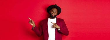 Kış tatili ve alışveriş konsepti. Şüpheci ve eğlendirilmemiş siyah adam parmaklarını sağa ve suratını ekşiten kırmızı arka planda duran, öneri önermeyen, hoşnutsuz.