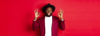 Noel alışverişi ve insanların konsepti. Tatmin olmuş Afro-Amerikan erkek model tamam işaretleri gösteriyor, gülümsüyor memnun, iyi seçimi övüyor, kırmızı arka plan.