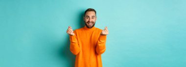 Gülümseyen yakışıklı adam kalp gösteriyor ve mutlu görünüyor, şanslı bir jest, açık mavi arka planda turuncu süveter içinde duruyor..
