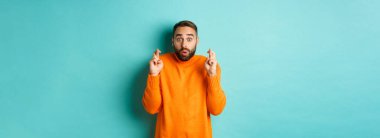 Umutlu adam heyecanla bekliyor, parmaklarını çapraz yapıp dilek tutuyor, turkuaz arka planda duruyor..