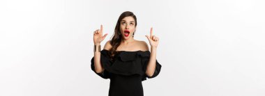 Moda ve güzellik. Siyah elbiseli, neşeli bir kadın beyaz arka planda dikilip reklam teklifini işaret ediyor..