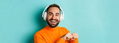 Kulaklıkla müzik dinleyen, turuncu süveter giymiş turkuaz arka planda duran yakışıklı modern adamın yakın çekimi..