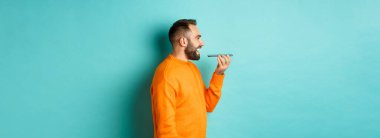 Yakışıklı sakallı bir adamın sesli mesaj kaydetme profili, cep telefonu tutmak ve hoparlörden konuşmak, mutlu bir şekilde gülümsemek, turkuaz arka planda durmak..