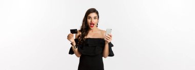 İnternetten alışveriş konsepti. Siyah elbiseli şık bir kadın, elinde akıllı telefonlu bir kredi kartı var, heyecanlı görünüyor, beyaz arka planda duruyor..