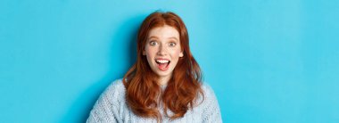 Şaşırmış ve heyecanlı kızıl saçlı kızın yakın çekimi inanılmaz haberler duyuyor, kameraya bakıyor, tanıtım teklifine bakıyor, mavi arka planda duruyor..