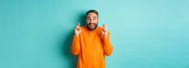 Heyecanlı adam beklenen bir şey bekliyor, parmaklarını çapraz yapıp gülümsüyor, dilek tutuyor, açık mavi arka planda duruyor..