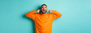 Hüsrana uğramış ve sinirli bir adam üst kattaki komşulara bağırıyor, parmaklarıyla kulaklarını kapatıyor, gürültülü rahatsız edici sesler çıkarıyor, açık mavi arka planda duruyor..