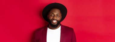 Sakallı, parti ceketi ve şık şapkalı, kameraya gülümseyen, kırmızı arka planlı yakışıklı Afro-Amerikan adam..