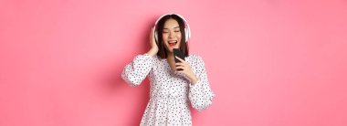 Tatlı Asyalı kadın karaoke uygulaması oynuyor, cep telefonuyla şarkı söylüyor, kulaklık takıyor, pembe arka plan üzerinde duruyor..