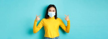 Covid-19, sosyal uzaklık ve salgın konsepti. Genç Asyalı kadın kendini koronavirüsten korur. Mavi arka planda duran tıbbi maskesini işaret eder..