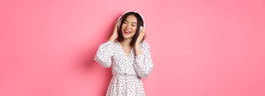Romantik Asyalı kadın mutlu gülümsüyor, kulaklıkla müzik dinliyor ve dans ediyor, pembe arka planda modaya uygun bir elbise giyiyor..