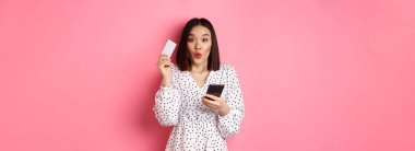 Çekici genç Asyalı kadın online sipariş veriyor, kredi kartı ve cep telefonu tutuyor, internet alışverişi yapıyor, pembe arka planda mutlu duruyor..