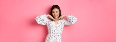 Moda elbiseli güzel Asyalı kadın sevimli bir yüz yapıyor, çene hattının yanında el ele tutuşuyor ve kameraya bakıp pembe arka planda dikiliyor..