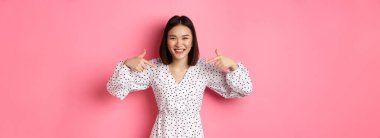 Güzel Koreli kadın logonuzu işaret ediyor ve gülümsüyor. Romantik pembe arka planda giyinmiş duruyor. Boşluğu kopyala