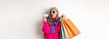 Modaya uygun Asyalı yaşlı kadın alışverişe gidiyor, elinde kağıt torbalar ve plastik kredi kartı var, şık güneş gözlüğü takıyor, beyaz arka planda duruyor..